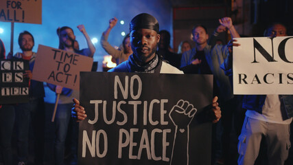 Portrait of black activist with No Justice No Peace placard among chanting crowd of protesters....