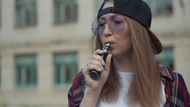 Close up pretty young hipster woman in black cap vape ecig. Girl vape popular ecig gadget, vaping device.Happy brunette vaper girl with e-cig. Portrait of smoker female model with electronic cigarette