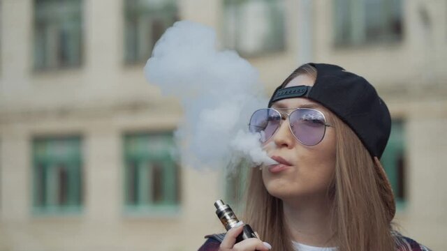Close up pretty young hipster woman in black cap vape ecig. Girl vape popular ecig gadget, vaping device.Happy brunette vaper girl with e-cig. Portrait of smoker female model with electronic cigarette