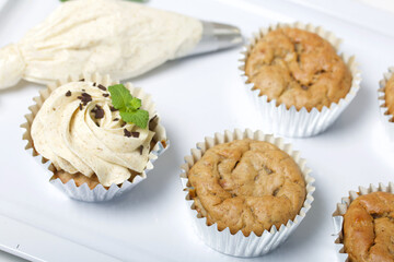 Banana cupcake. With butter cream and banana jam. Decorated with mint leaves and chocolate sprinkles. Next to them are cupcakes without cream and a pastry bag with cream.