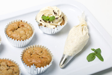 Banana cupcake. With butter cream and banana jam. Decorated with mint leaves and chocolate sprinkles. Next to them are cupcakes without cream and a pastry bag with cream.