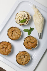Banana cupcake. With butter cream and banana jam. Decorated with mint leaves and chocolate sprinkles. Next to them are cupcakes without cream and a pastry bag with cream.