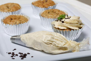 Banana cupcake. With butter cream and banana jam. Decorated with mint leaves and chocolate sprinkles. Next to them are cupcakes without cream and a pastry bag with cream.