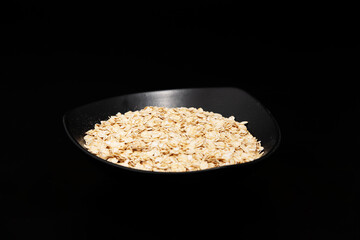 Raw dry oat flakes cereal heap in black ceramic bowl top view close up isolated