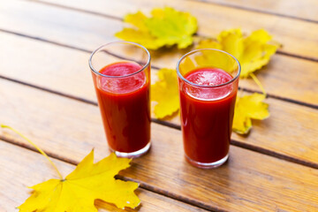 Tomato juice is poured into two glass glasses on a wooden table with yellow fallen maple leaves, in autumn. Selective focus.