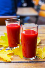 Tomato juice is poured into two glass glasses on a wooden table with yellow fallen maple leaves, in autumn. Selective focus.