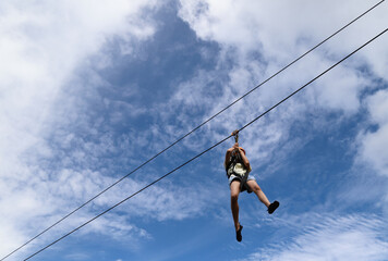 Closeup of a boy gliding on extreme trolley zip-line in adventure park with cloudy blue sky background. 