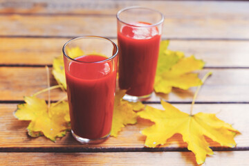 Tomato juice is poured into two glass glasses on a wooden table with yellow fallen maple leaves, in autumn. Selective focus. Tinted background