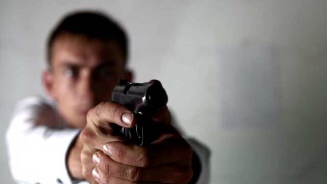 Man in white shirt is raising his hands with a gun and taking aim, preparing to fire in slow motion. He is standing on white blur background holding a handgun before him.