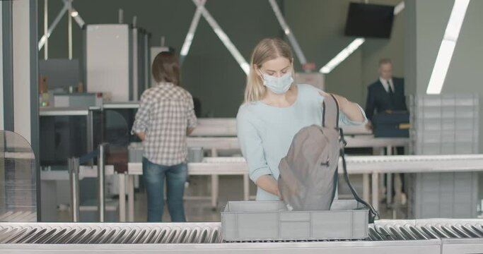 Woman In Face Mask Putting Personal Belongings In Tray And Passing Security Gates Of Airport. People Check-in At The Background In Departure Terminal. Tourism On Covid-19 Pandemic. Cinema 4k ProRes HQ