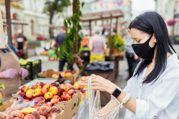 Portrait of pretty house wife in protective mask holding net bag and buying organic products at farmers market during coronavirus. New social rules after pandemic.