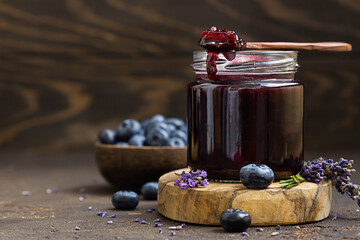 Glass jar and spoon with delicious blueberry jam with fresh berries, lavender and rosemary on brown...