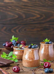 Portion jars with chocolate mousse or pudding decorated with sweet cherry, blueberry and mint and pieces of dark chocolate. Selective focus. Rustic style. Dark background.
