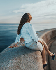A young unrecognizable fashionable girl sitting at the edge of the shore watching the sunset over the sea.
