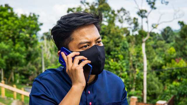 A portrait of Asian Malay man with Baju Melayu cloth wearing a black fabric face mask talking on the phone. Hygiene lifestyle. New normal concept.