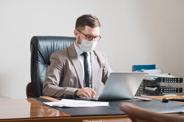 Young businessman in glasses and face mask working on a laptop in his personal office. Business during quarantine.