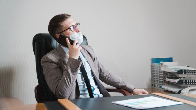 Young businessman in glasses and face mask talking to a smartphone in his personal office. Business during quarantine.