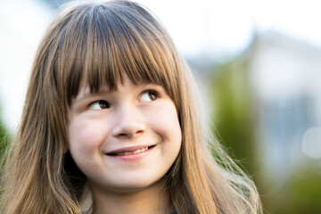 Fototapeta premium Portrait of pretty child girl with gray eyes and long fair hair smiling outdoors on blurred green bright background. Cute female kid on warm summer day outside.