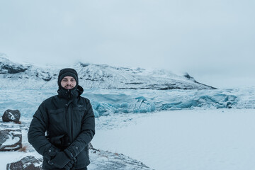 Fototapeta na wymiar A man is standing in front of a glacier with plenty of snow surrounding him. He is dressed for cold conditions and the day is gloomy