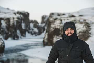 Fototapeta na wymiar A man standing in front of a gorge with a river flowing behind him. There is plenty of snow around and the man is dressed for cold weather