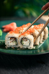 Took a sushi roll by the chopsticks. The range of different types of sushi, rolls and Maki with sauces and chopsticks. On black rustic background