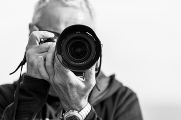 A black and white photo of a middle-aged man, in his 50th, takes a picture with a modern mirrorless camera. The camera covers his face. Blue hazy sky in the background