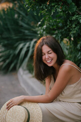 Young beautiful woman wearing straw hat and linen beige suit posing near bush of blooming roses at sunset.