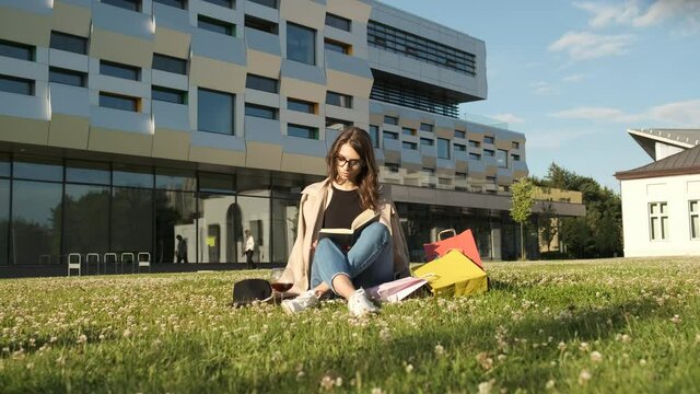 Stylish girl in glasses and a brown cloak sits in the park on the grass and reads a book and smiles at the camera. Gift packages. Sunny summer evening