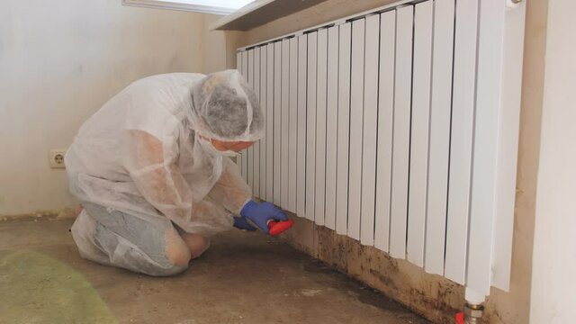 A woman in a protective suit and a respirator sprays a special antifungal spray on the mold-infested wall under the heating battery.