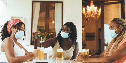 Three young African American girlfriends sitting at brewery (pub) with beer glasses wearing...