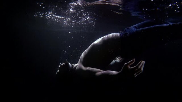 Close-up of a bearded man falling back into the dark water, he is in his pants and with a naked torso. There is darkness and bubbles around it, and a little light comes from the surface of the water