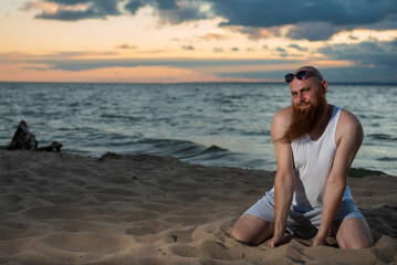 Fototapeta na wymiar Funny bald man with red beard posing on the beach at sunset. A humorous male parody of a glamorous girl.