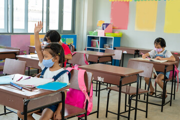 Portrait of an school kids wearing protective mask  raising hands to answer in an elementary school class.