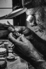 A black and white photo of a watchmaker examining the balance wheel of a watch using magnifying glass under a an electric lamp.