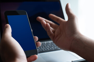 Mockup man using blue screen mobile smart phone while working on laptop computer at office