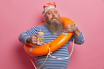 Portrait of bearded stout sailor with lifebuoy, drinks refreshing water, looks thoughtfully away,...