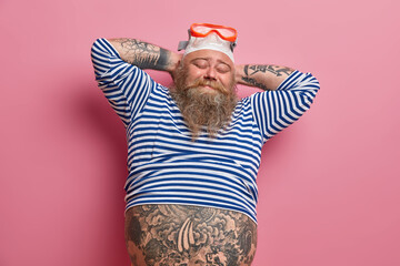 Relaxed tattooed male swimmer keeps hands behind head, closes eyes, wears swimming goggles and cap,...