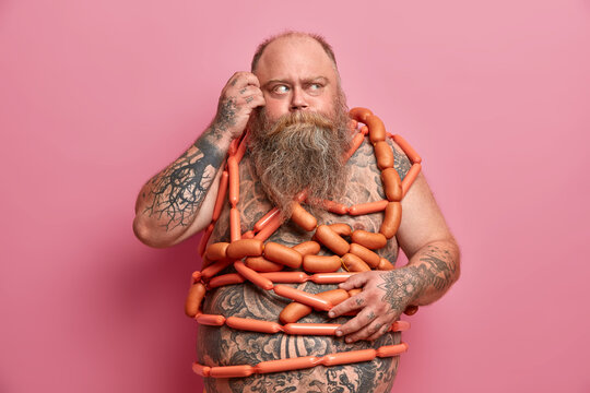 Thick Bearded Man Wrapped In Sausages, Scratches Head And Looks Away Thoughtfully, Obsessed With Food, Leads Unhealthy Lifestyle, Isolated On Pink. People, Obesity, Diet And Weight Loss Concept