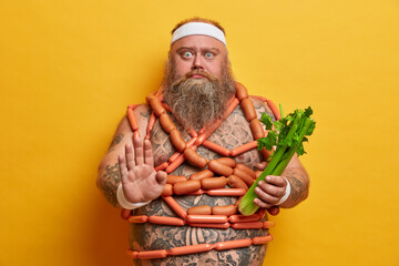 Serious motivated overweight man makes stop gesture, keeps to strict vegetarian diet and chooses celery to eat, wears sport headband, stands naked, wrapped by sausages, stands indoor. Junk food - obrazy, fototapety, plakaty