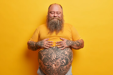 Pleased stout man keeps hands on belly, feels satiety after eating delicious dinner, stands with...