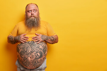 Photo of overweight pensive man keeps hands on big belly with tattoo, thinks and looks aside, has...
