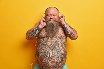 Optimistic carefree naked man with big belly, enjoys awesome beat in stereo headphones, closes eyes, has problem of overweight, isolated on yellow background. Listening new track on music platform