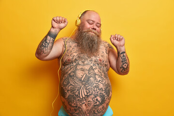 Happy bare man with fat stomach, tattooed belly, enjoys listening new song in headphones, raises arms, clenches fists, moves with rthythm, feels carefree, enjoys fantastic bits, poses indoor - Powered by Adobe
