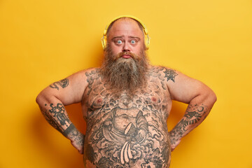 Photo of sad plump bearded man looks with surprised expression at camera, listens favorite song in...