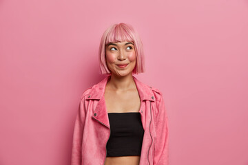 Studio shot of fashionable woman has bobbed pink hair and fringe, looks with happy dreamy expression, tempting look up, wears black top with stylish rosy jacket. Pleasant emotions, style concept