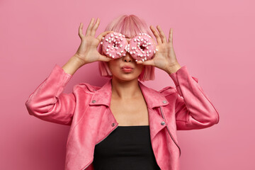 Fashionable woman has pink hairstyle, covers eyes with two delicious doughnuts, has fun and eats junk food, wears stylish jacket, poses over rosy wall. Tasty dessert. Going to eat appetizing food