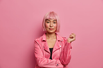 Serious stylish lady looks with calm expression direcrly at camera, has healthy skin, rouge cheeks, wears pink hair wig, dressed in jacket, keeps hand raised, isolated over rosy background. Monochrome