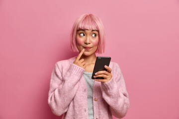 Stylish young woman has dyed rosy hair, holds smartphone, looks away with pensive expression, wears sweater, thinks what answer to give on received message, wears soft pink jumper, stands indoor