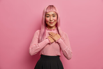 Young attractive grateful woman with pink hair clasps hands to heart, shows heartleft gratitude,...