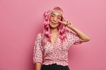 Happy pink haired girl applies eye patches for reducing dark circles, makes peace sign, wears hair...